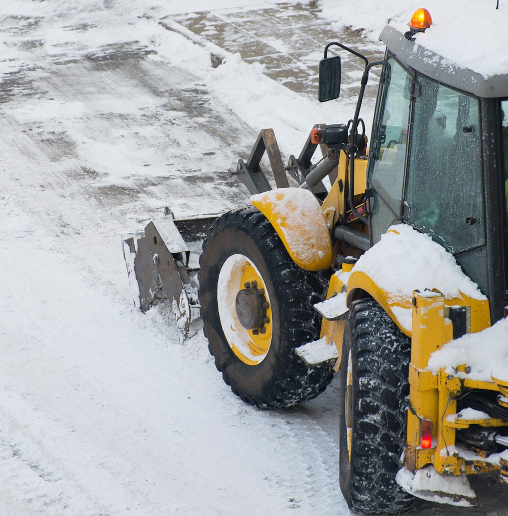 A yellow snowplow clears snow from a concrete path during winter, leaving a clean surface behind. The machine's top light is on.