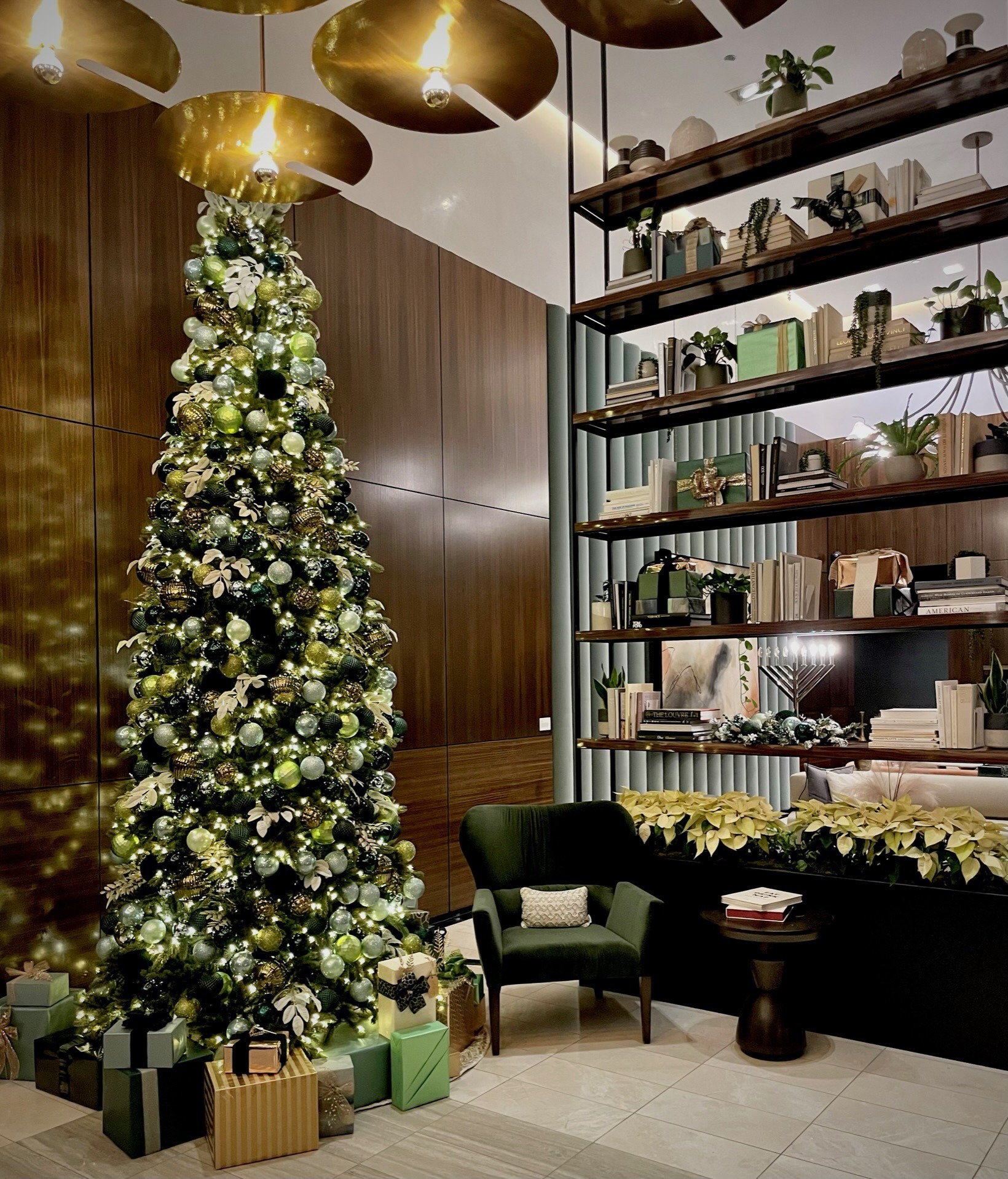 A tall, decorated Christmas tree stands in a modern, cozy room with bookshelves, plants, and a comfortable chair. Gifts surround the tree.