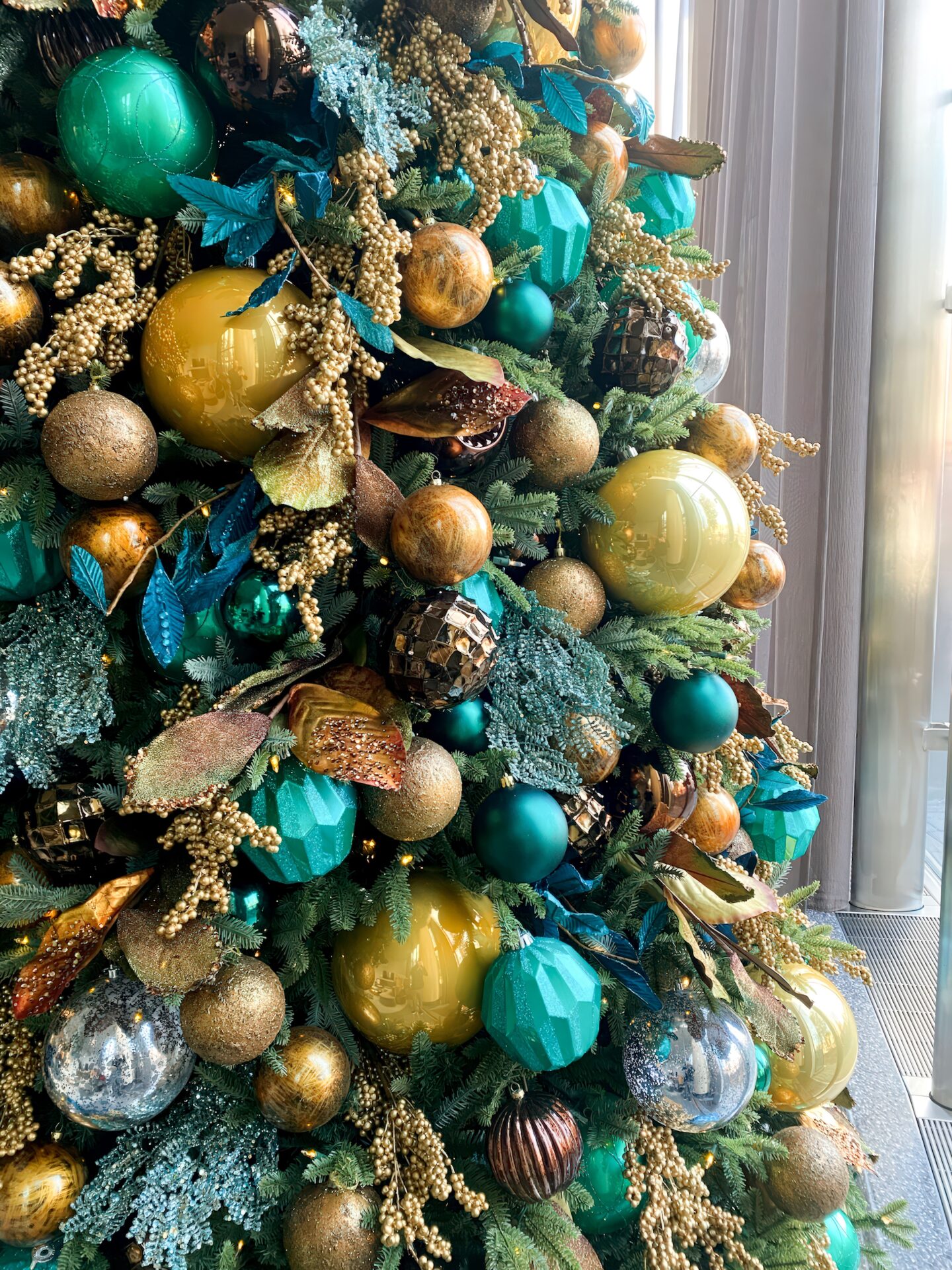 A colorful Christmas tree with green, gold, and bronze ornaments, adorned with artificial leaves and berries, set indoors near a glass window.